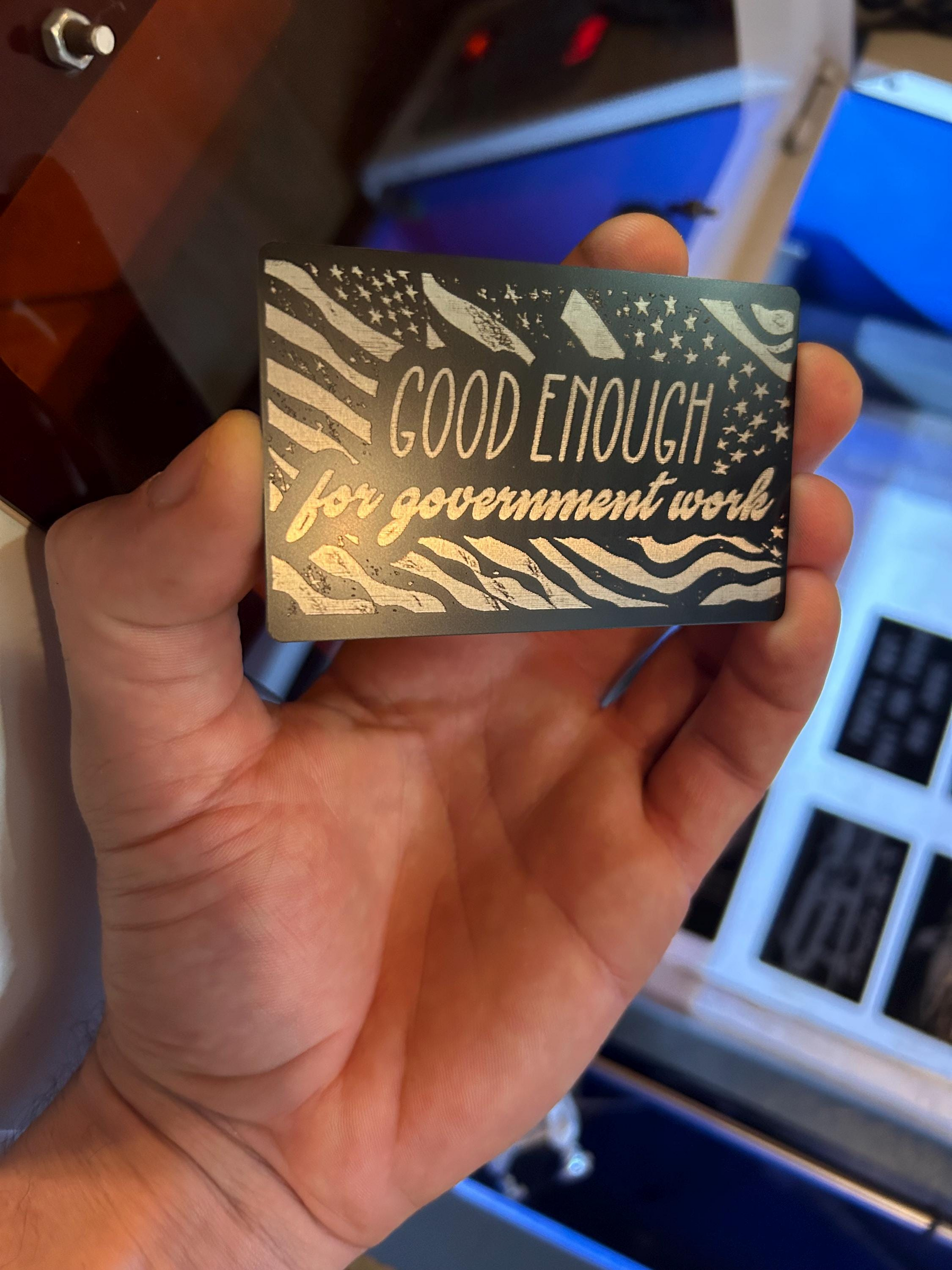 Hand holding a metal novelty card with 'Good Enough for Government Work' text in front of a laser engraving