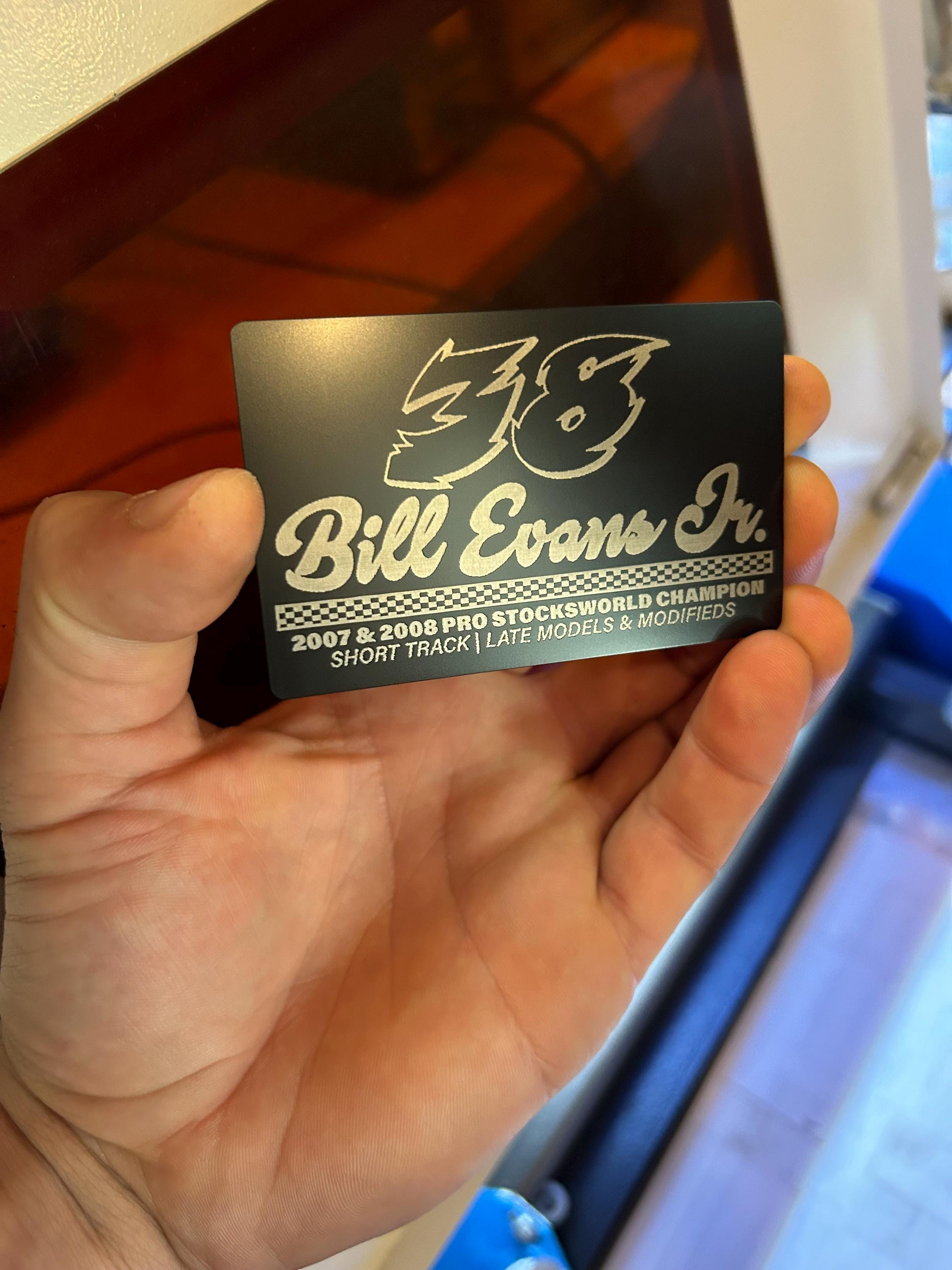 Hand holding a black/silver metal business card with stock car racing driver personalized layout design, in front of a laser machine