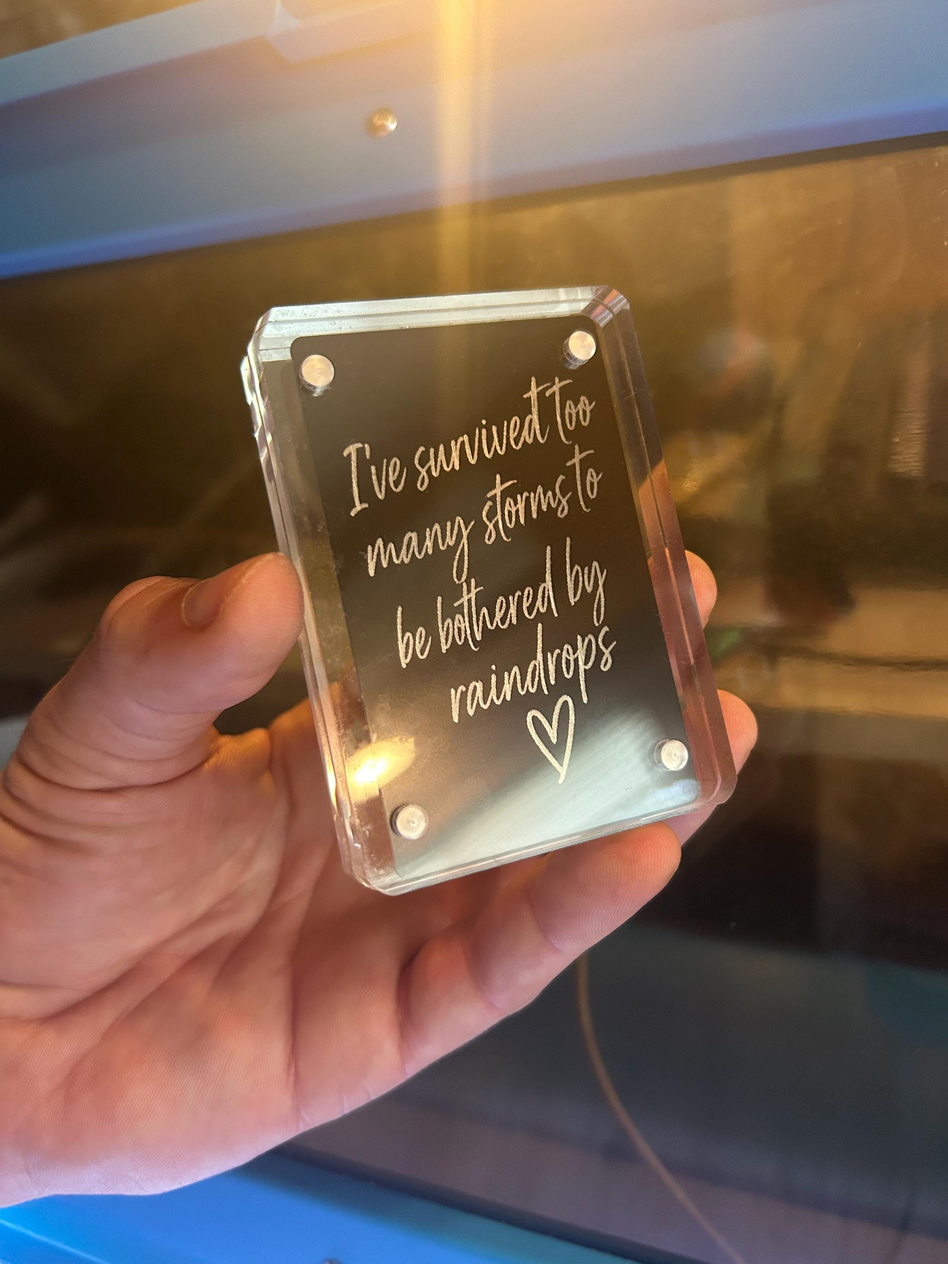 Hand presenting a metallic black and silver inspirational card engraved with rainstorm resilience message and cute heart design