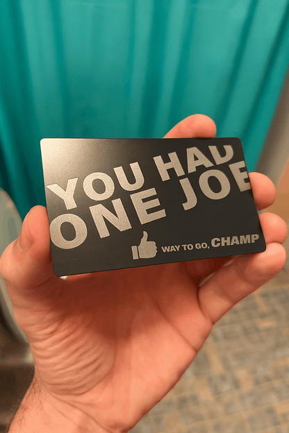 Hand holding a metal gag gift card with humorous You Had One Job text against a teal curtain background