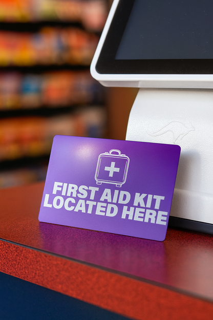 Purple sign indicating a first aid kit location on a checkout counter with a blurred store background.