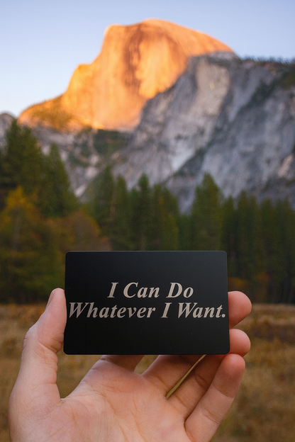 Hand holding a metal card with 'I Can Do Whatever I Want' text in front of a mountain landscape.