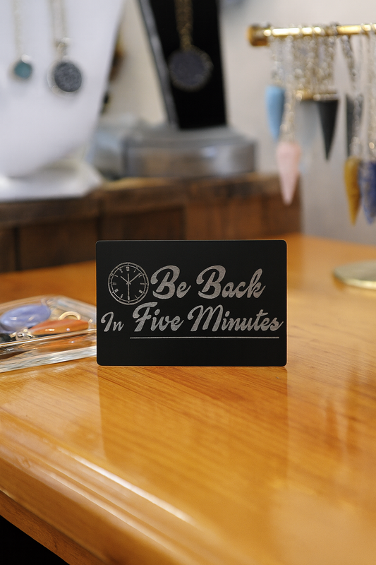 Black metal signage with 'Be Back In Five Minutes' text on a wooden surface with jewelry in the background