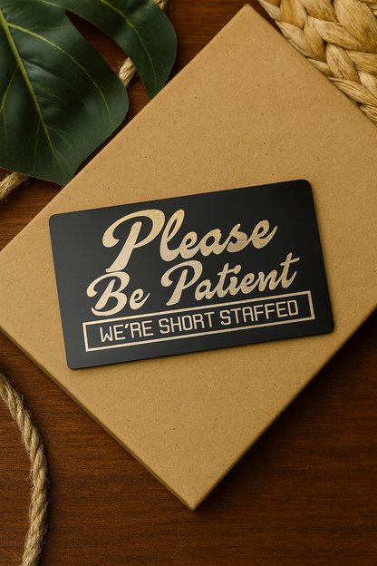 Black/silver metal sign with 'Please Be Patient, We're Short Staffed' text on a brown paper bag.