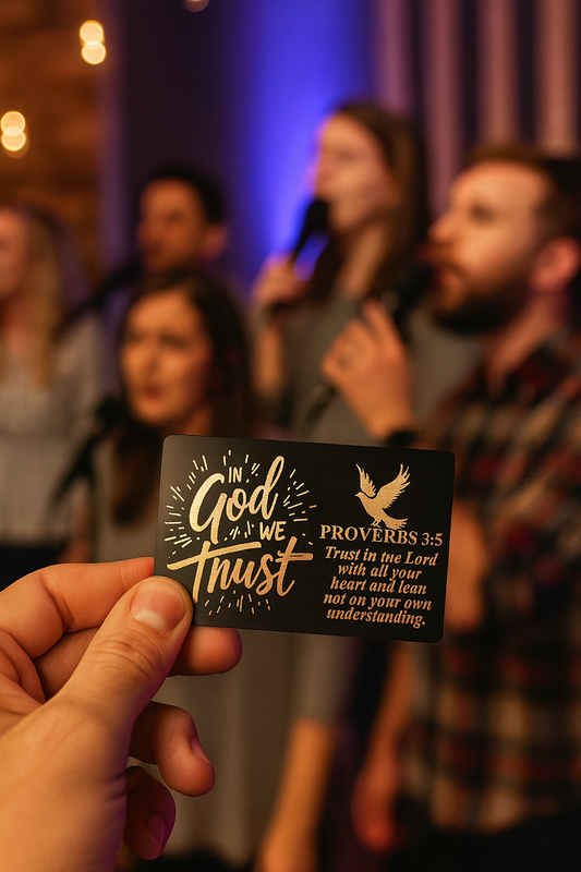 Hand holding a card with 'In God We Trust' proverbs 3:5 text in a blurred praise and worship setting
