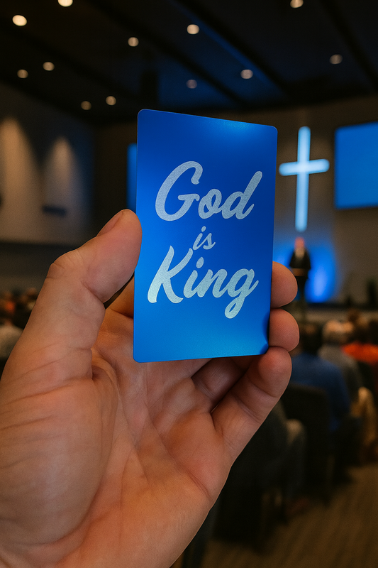Hand holding a blue/silver metal engraved card with 'God is King' text in a blurred church setting