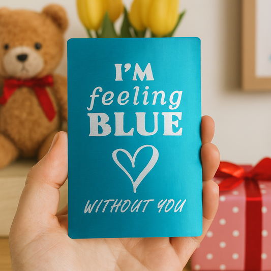 Hand holding a blue card with 'I'm feeling BLUE without you' text, surrounded by a teddy bear, yellow tulips, and a gift.