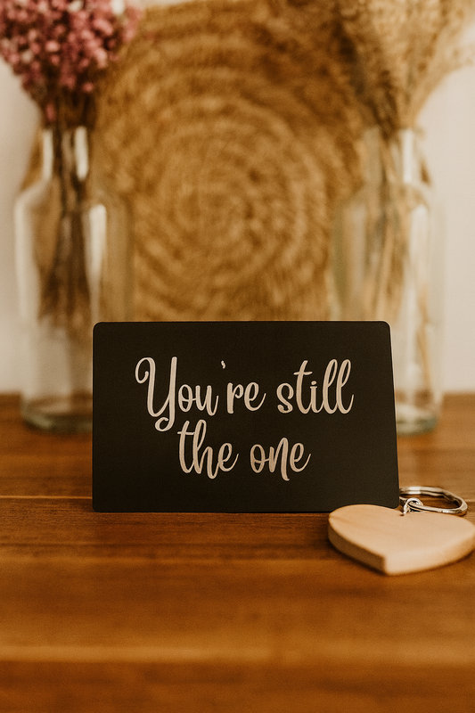 Black/silver metal laser engraved mini sign with 'You're still the one' text on a wooden surface with a heart-shaped object.