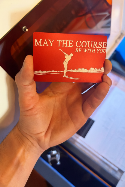 Hand holding a red/silver metal card with a golf design and inspirational text, in an industrial machine setting