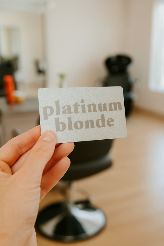 Hand holding a silver metal card with 'platinum blonde' text in a blurred hair salon setting