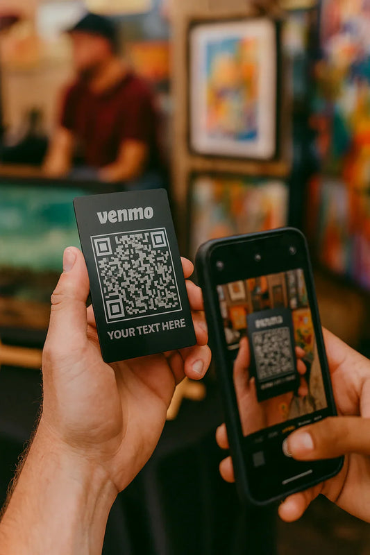 Two hands holding a Venmo QR code card and a smartphone with a blurred background of an artist festival market.