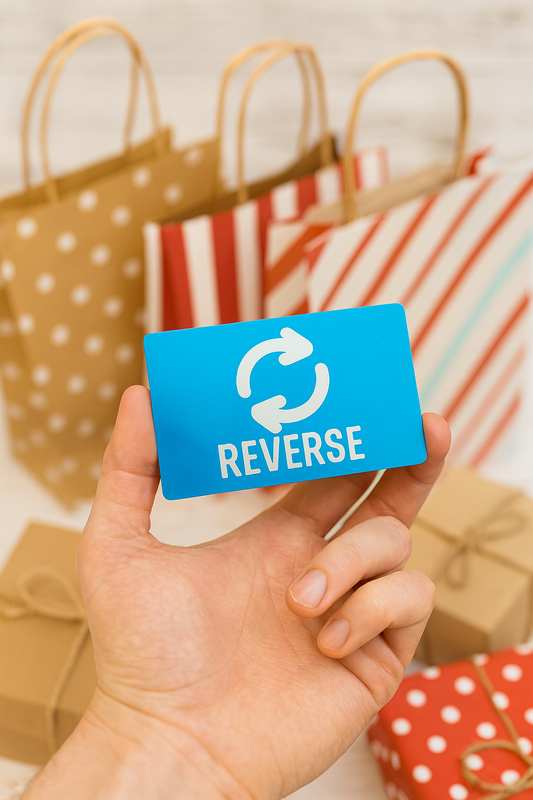 Hand holding a blue/silver laser engraved gift card with 'REVERSE' and a circular arrow symbol, with gift bags in the background.