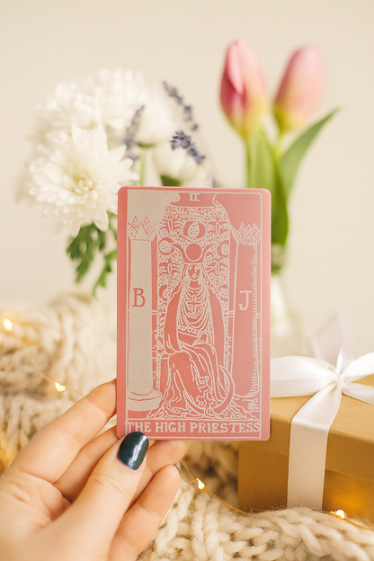 Rose gold metal tarot card held by a hand with flowers and gift in the background