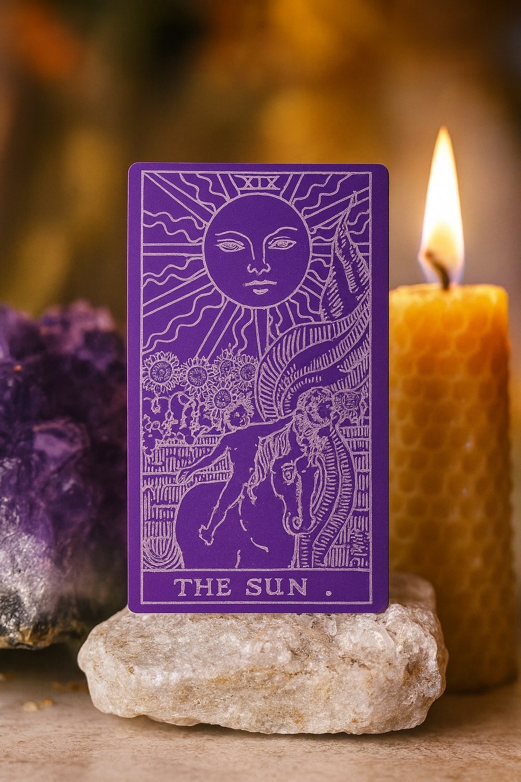 Purple/silver metal tarot card titled 'The Sun' with a sun and moon design, placed on a rock with a candle and crystal in the background.