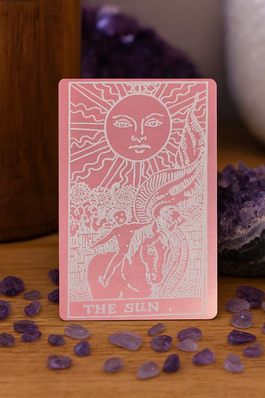 Rose Gold/ Silver laser engraved metal tarot card titled 'The Sun' with a sun and rider illustration, surrounded by purple crystals on a wooden surface.