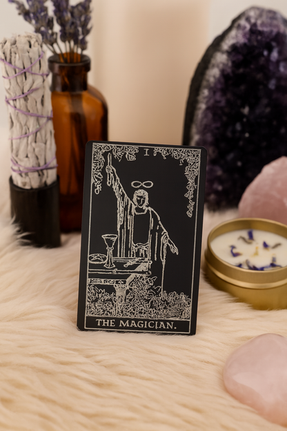 Metal tarot card titled 'The Magician' surrounded by mystical items like a crystal and candles on a soft surface.