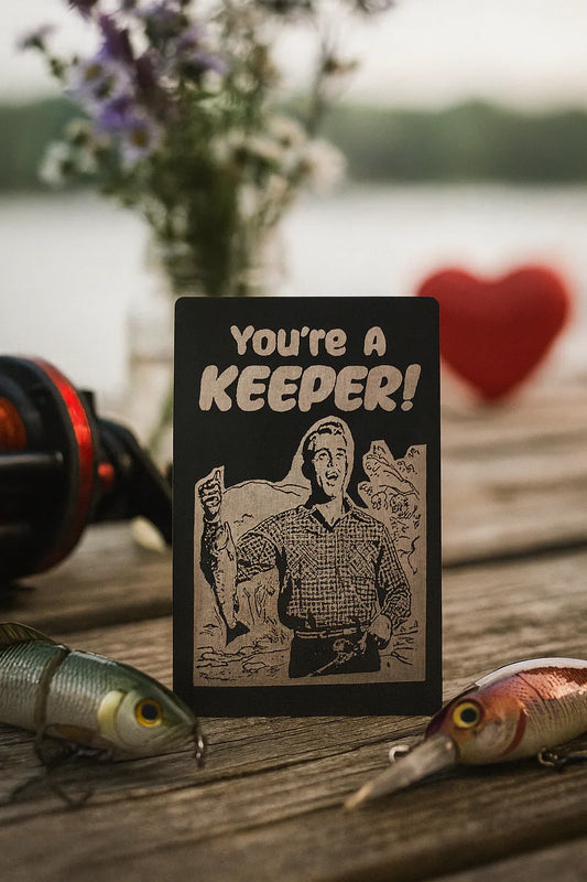 Card with 'You're a Keeper!' text on a wooden surface with fishing equipment and flowers.