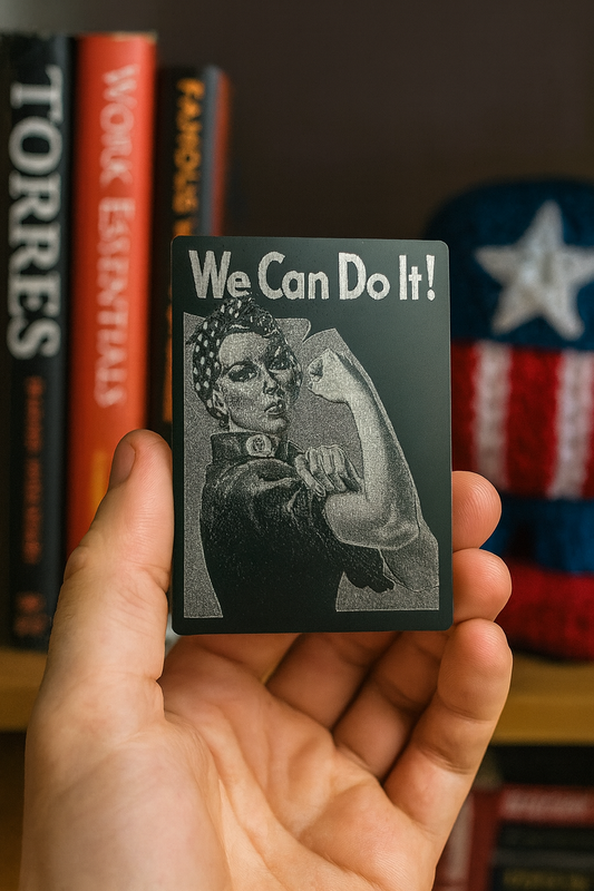 Hand holding a metal laser engraved inspirational card with 'We Can Do It!' text and a Rosie the Riveter graphic, with books and Americna art in the background.