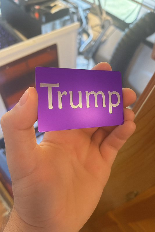 Hand holding a purple metal card with 'Trump' laser engraved on it, blurred background