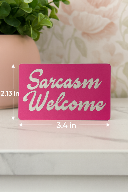 Pink sign with 'Sarcasm Welcome' text and measurements on a white surface with decorative elements.