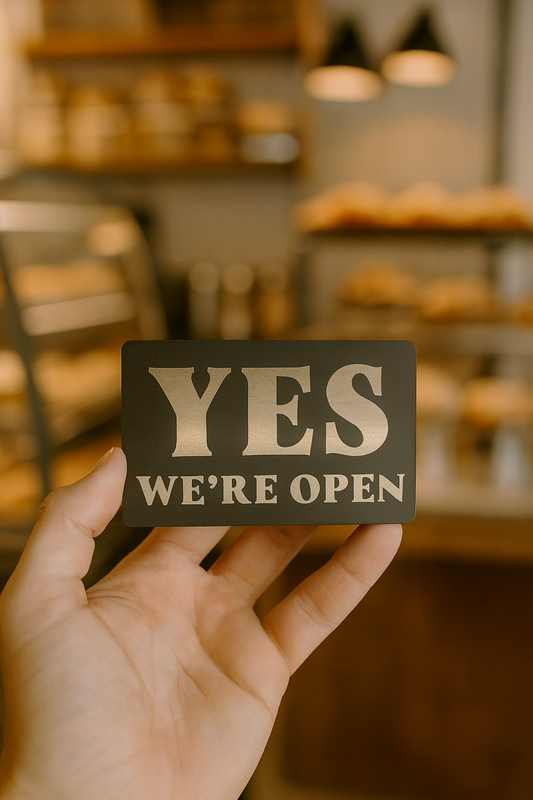 Hand holding a 'YES WE'RE OPEN' sign in front of a blurred bakery background
