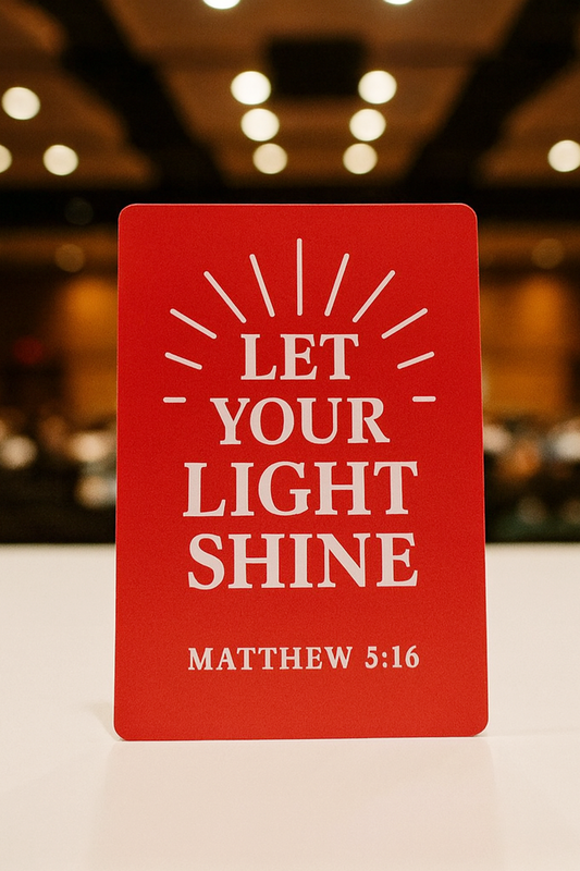 Red/silver metal bookmark card with 'Let Your Light Shine' text on a blurred indoor background