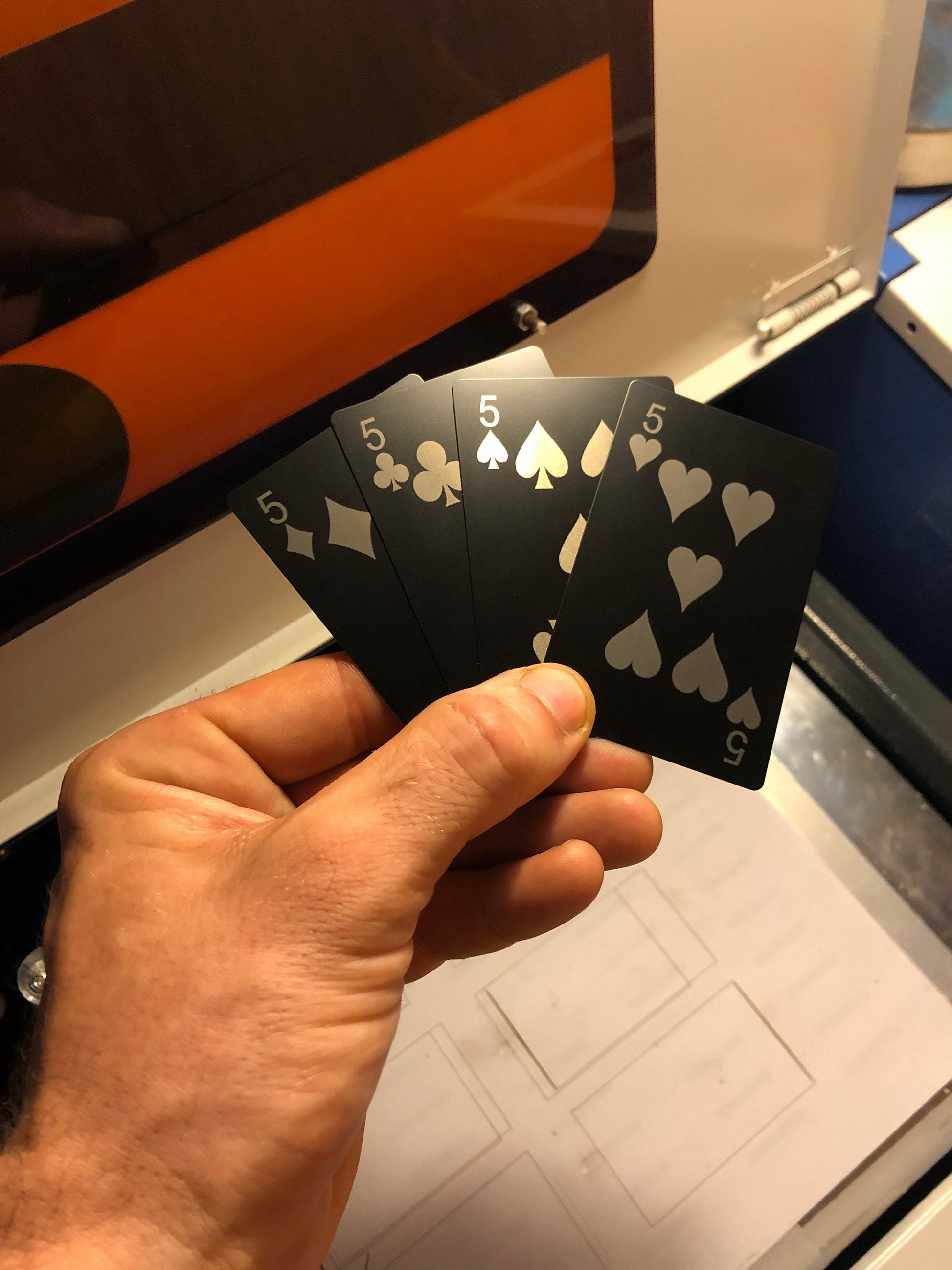 Hand holding a deck of metal playing cards, wth Five of Diamonds, Five of Clubs, Five of Spades, Five of Hearts laser engraved in matte black/silver anodized aluminum