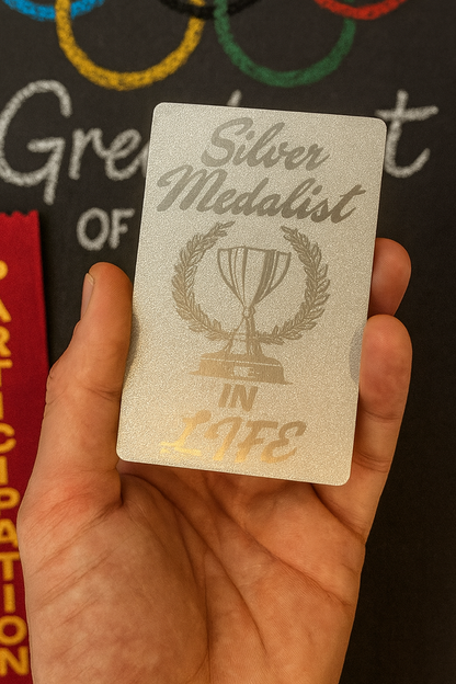 Pearl silver metal laser engraved gag gift card with 'Silver Medalist in Life' design held in a hand against a blurred sports background
