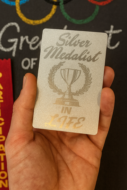 Pearl silver metal laser engraved gag gift card with 'Silver Medalist in Life' design held in a hand against a blurred sports background