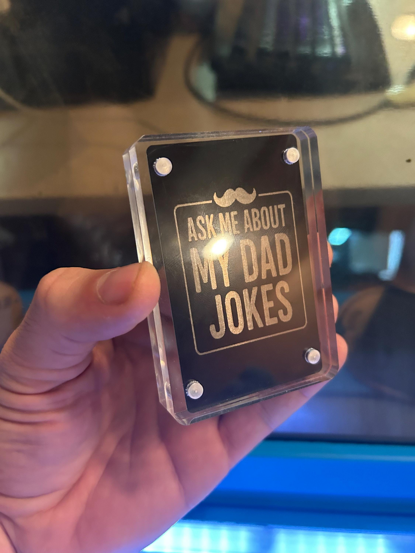 Hand holding a laser engraved black and silver metal gag gift card that reads “Ask Me About My Dad Jokes” with mustache design