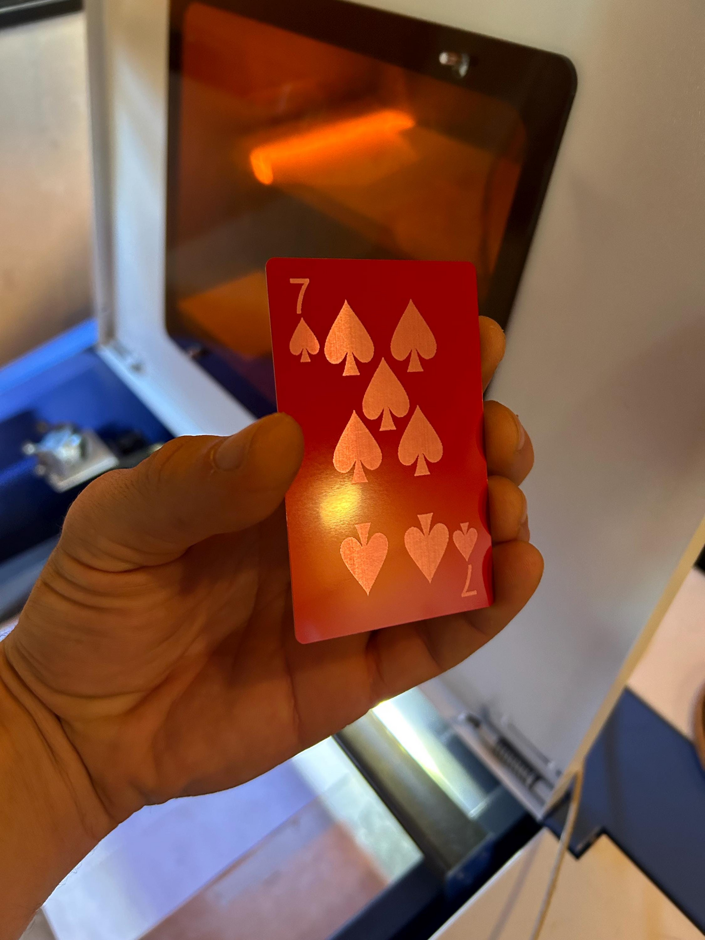 Hand holding a red and silver laser engraved playing card with a number 7 and spade symbols in front of a machine.