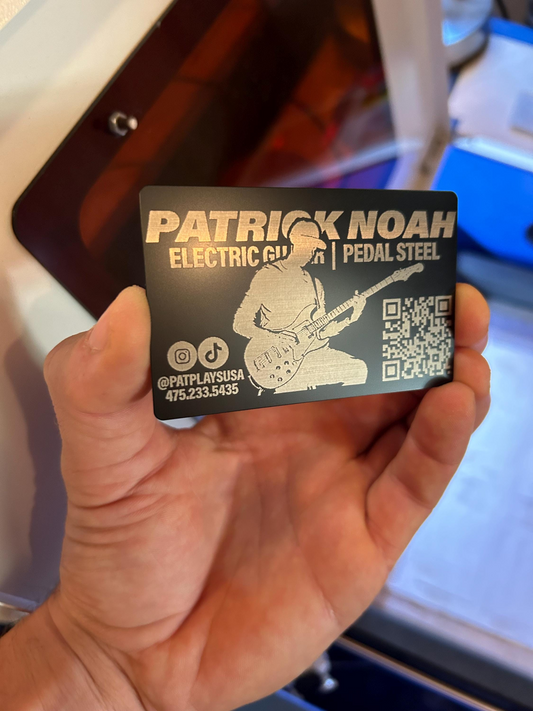 Hand holding a metal business card in black/silver with a pro guitarist engraving with personalized image text and contact details, shown in front of a laser engraving machine