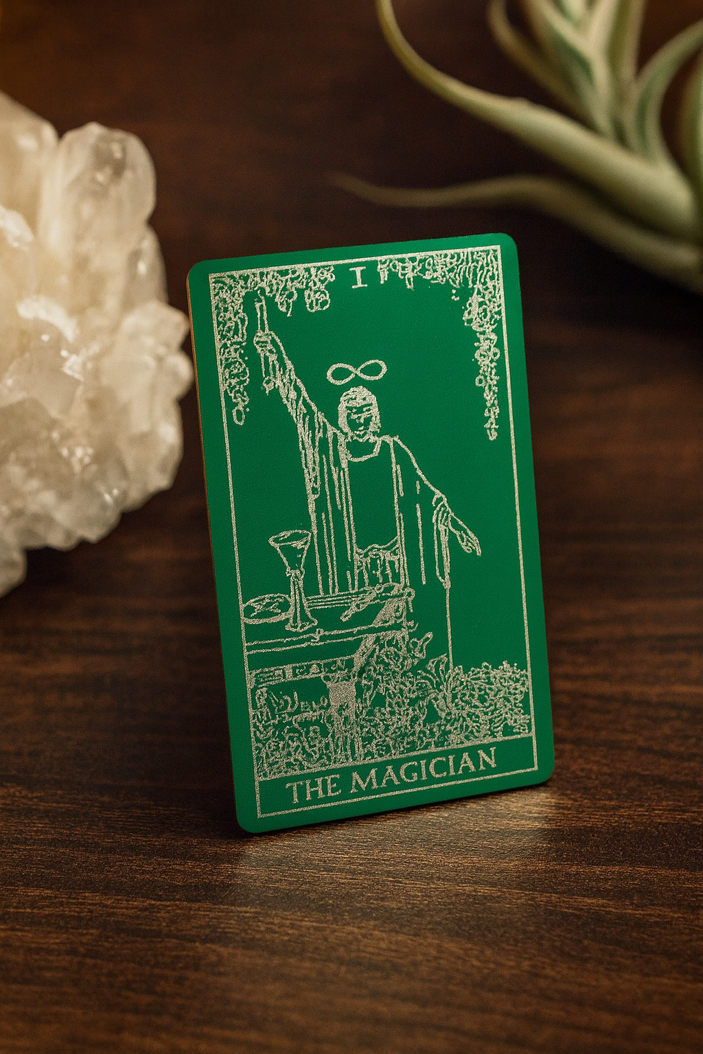 Green and silver metal tarot card titled 'The Magician' engraving on a wooden surface with crystals and plants.