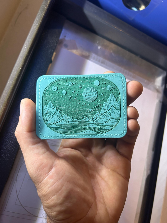 Teal engraved leather patch with celestial galaxy alien planet design held in a hand