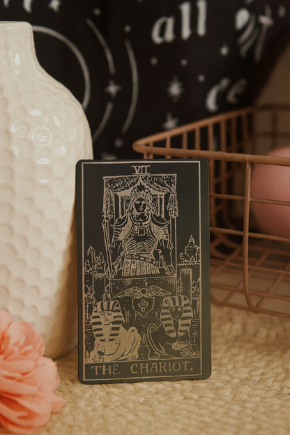 Metal tarot card titled 'The Chariot' on a textured surface with decorative items in the background.