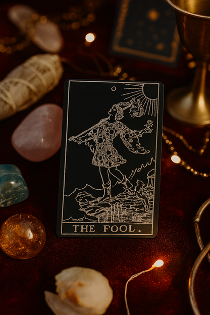 Metal tarot card titled 'The Fool' surrounded by crystals and other mystical items on a dark surface.