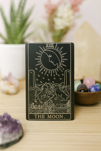 Metal black/silver Tarot card titled 'The Moon' on a wooden surface with decorative items.