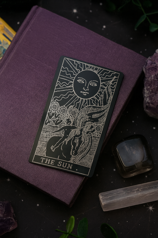 Purple book with a metal tarot card titled 'The Sun' on a dark background with crystals and plants.