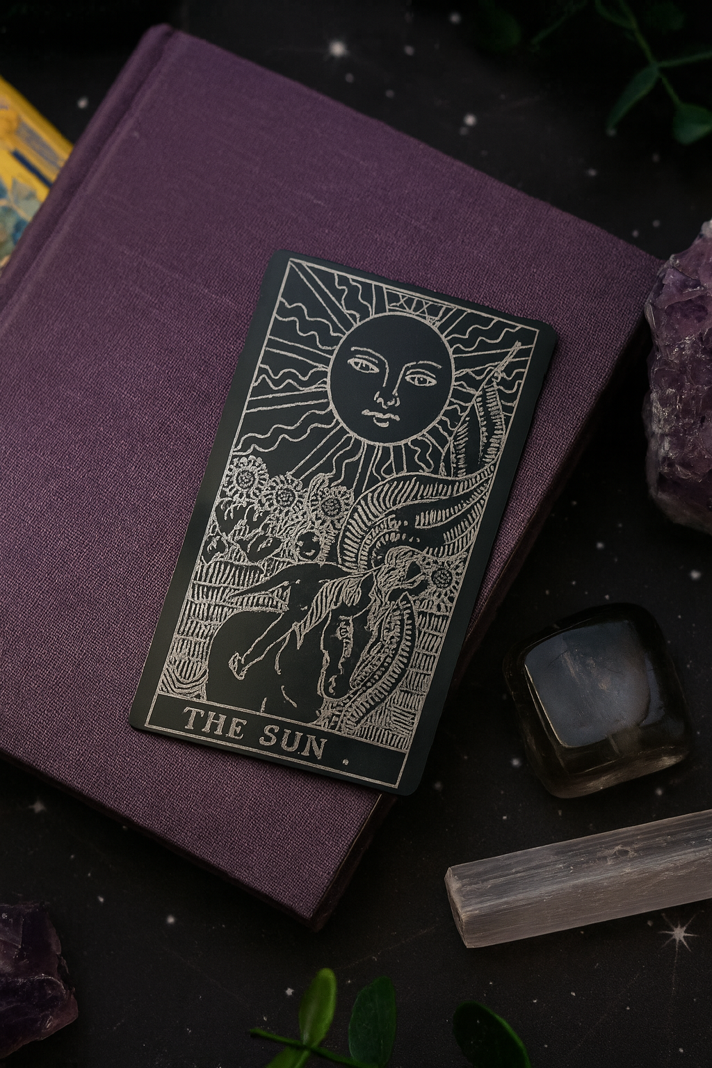 Purple book with a metal tarot card titled 'The Sun' on a dark background with crystals and plants.