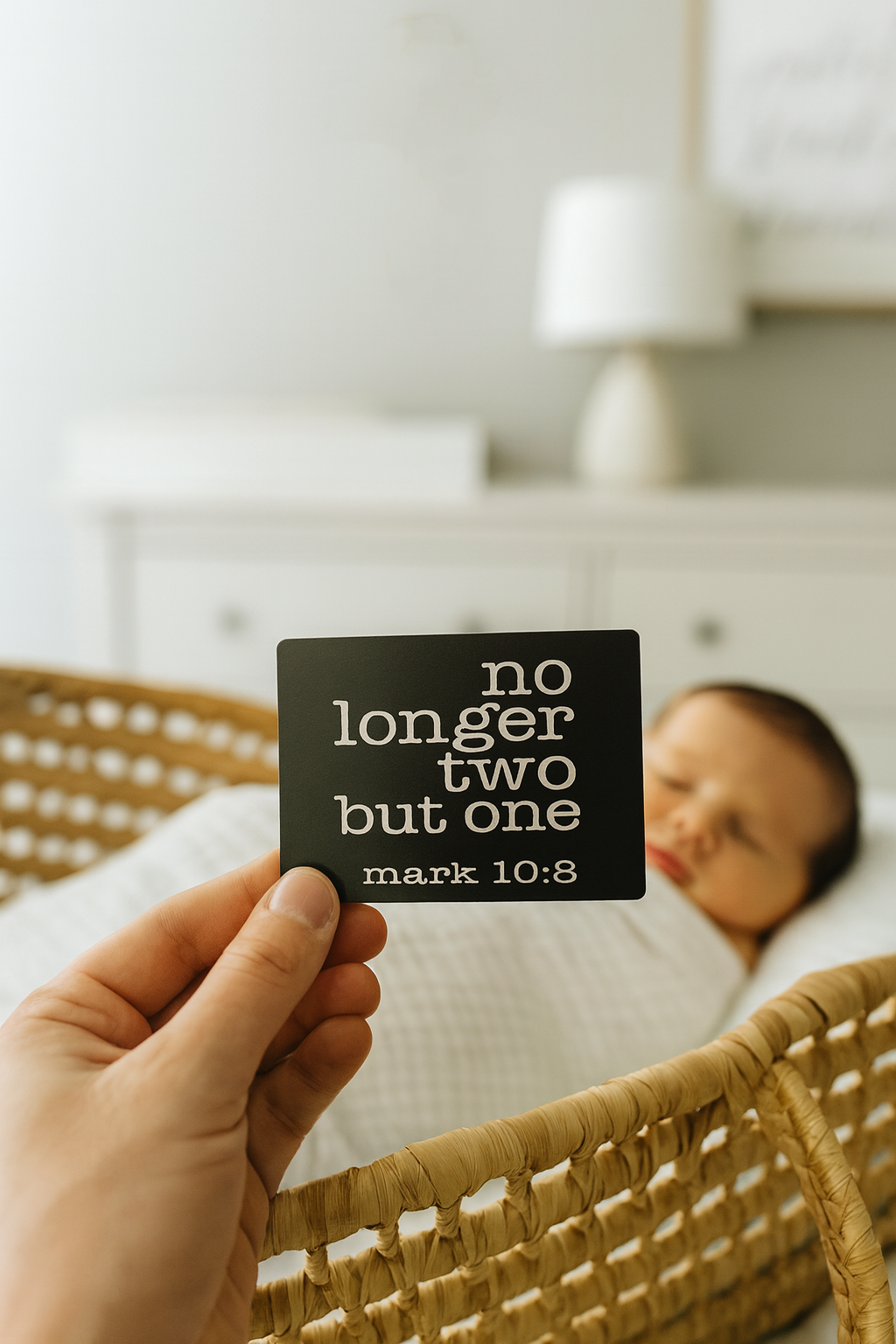 Person holding a laser engraved metal business card with 'no longer two but one' and mark 10:8 bible verse reference text in a bedroom setting with newborn baby inbackground.