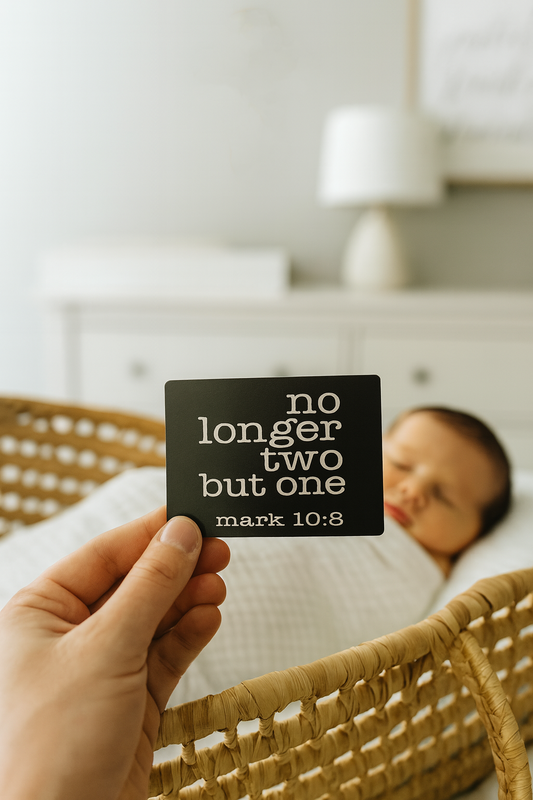 Person holding a laser engraved metal business card with 'no longer two but one' and mark 10:8 bible verse reference text in a bedroom setting with newborn baby inbackground.