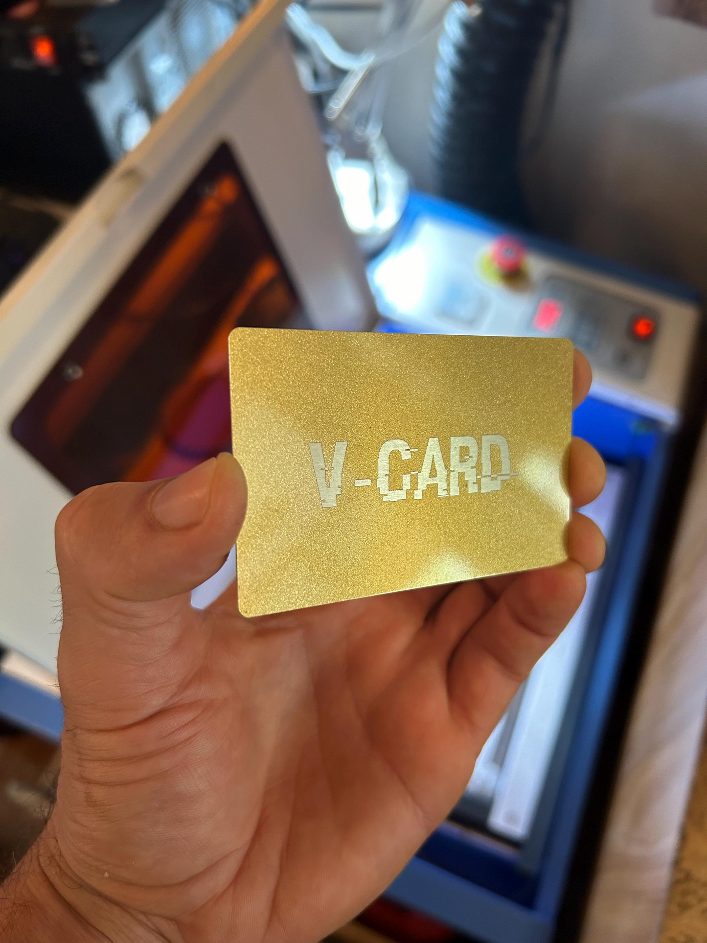 Hand holding a gold over silver metal business card, with personalized text engraving in front of laser cutting machine