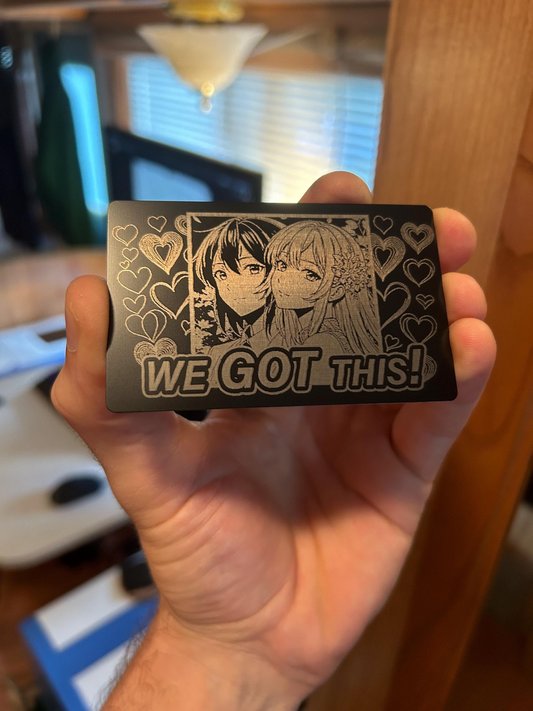 Metal card with laser engraved illustration of two female manga art protagonists and 'WE GOT THIS!' text, held in a hand.