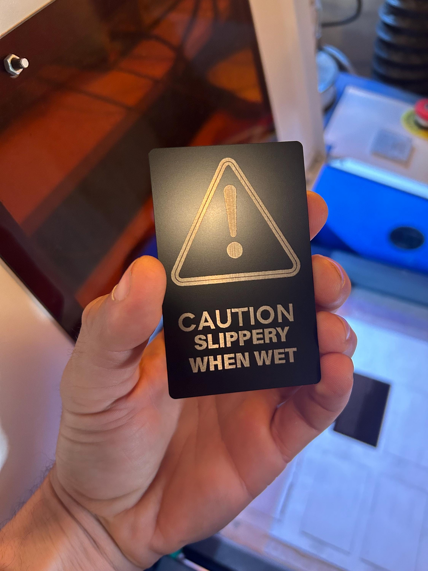 Hand holding a laser engraved metal retail warning sign with a warning triangle and text 'Caution Slippery When Wet' in an indoor setting.