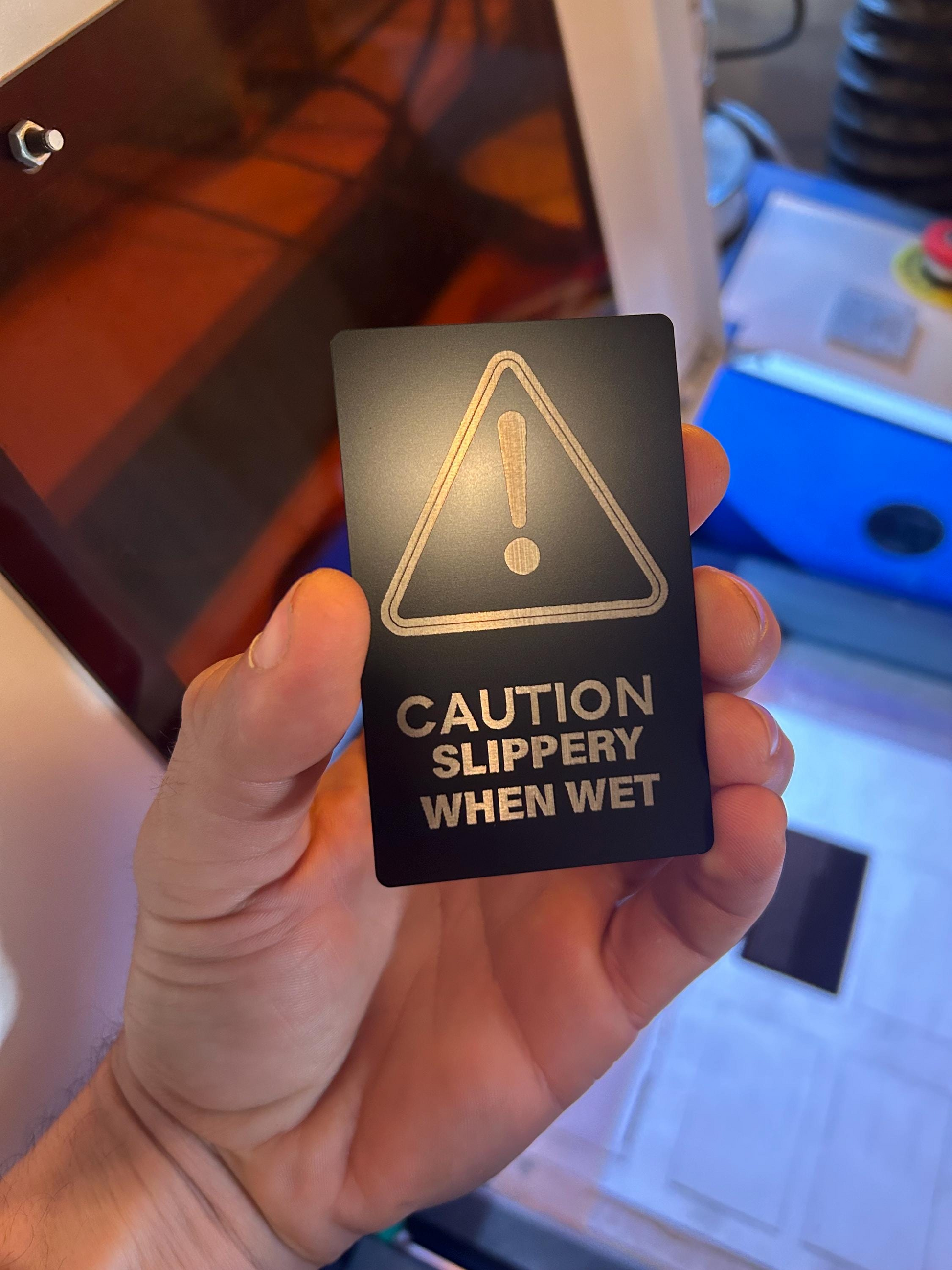 Hand holding a laser engraved metal retail warning sign with a warning triangle and text 'Caution Slippery When Wet' in an indoor setting.